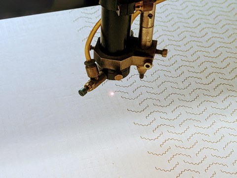 Das Bild zeigt einseitige Wellpappe, die mit dem Liner nach oben in einer Laserbearbeitungsmaschine liegt. Der Laser bringt gerade ein wellenförmiges Muster in die Wellpappe ein, um deren Formarbeit zu erhöhen.