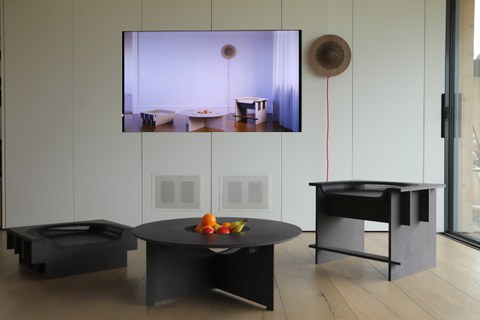 Im Bild ist im Vordergrund eine schwarze Sitzgruppe mit Tisch zu sehen, deren Oberflächen aus 3D umgeformten Holzwerkstoffen bestehen. Im Hintergrund ist eine Wandlampe aus dem gleichen Material zu sehen und daneben ein Bildschirm mit dem Entwurf.
