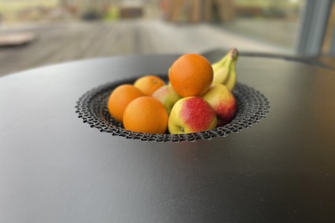 Das Bild zeigt eine Detailaufnahme eines Tisches mit mittig ausgeformter Vertiefung aus einem partiell perforierten Holzwerkstoff. In der Vertiefung liegt Obst.