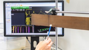 Das Bild zeigt einen Messaufbau zur Modalanalyse von Holzproben. Im Vordergrund wird eine frei hängende Holzprobe mit einem Impulshammer per Hand angeschlagen. Im Hintergrund ist ein Bildschirm mit der Software zur Auswertung des Versuches zu sehen.