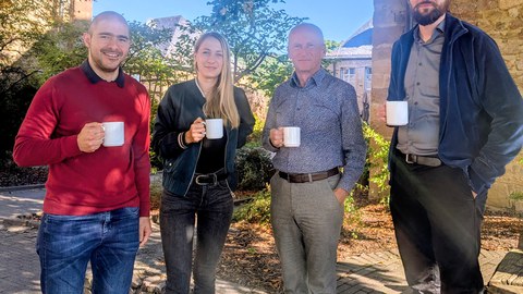 Drei Doktorand:innen und ein Professor stehen mit Kaffetassen am Rande einer Konferenz. 