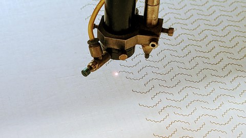 Das Bild zeigt einseitige Wellpappe, die mit dem Liner nach oben in einer Laserbearbeitungsmaschine liegt. Der Laser bringt gerade ein wellenförmiges Muster in die Wellpappe ein, um deren Formarbeit zu erhöhen.