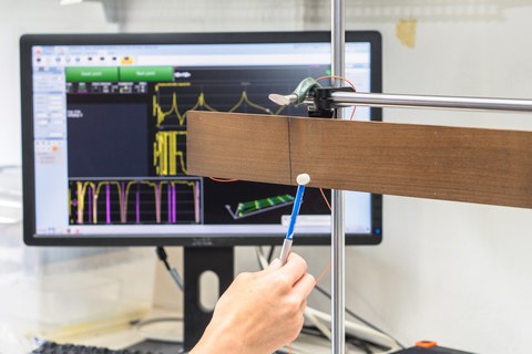 Das Bild zeigt einen Messaufbau zur Modalanalyse von Holzproben. Im Vordergrund wird eine frei hängende Holzprobe mit einem Impulshammer per Hand angeschlagen. Im Hintergrund ist ein Bildschirm mit der Software zur Auswertung des Versuches zu sehen.