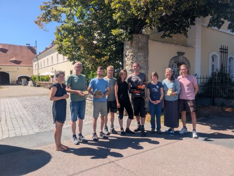 Das Bild zeigt das Projektkonsortium von MicroGrow: 4 Frauen und 5 Männer. Sie stehen vor dem Hof Podemus - einem gelb/weißen 3-Seitenhof mit vielen Sandsteinelementen.
