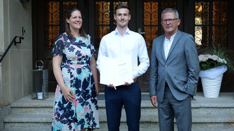 Das Bild zeit den Preisträger, Julian Sachsenweger, vor einem eisernen Tor auf einer Treppe mit der Urkunde des Preises. Rechts und links von ihm stehen die Preisverleiher, ein Mann und eine Frau.. 
