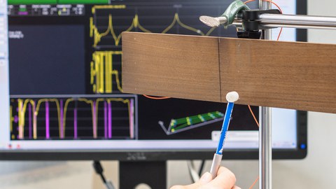 Das Bild zeigt einen Messaufbau zur Modalanalyse von Holzproben. Im Vordergrund wird eine frei hängende Holzprobe mit einem Impulshammer per Hand angeschlagen. Im Hintergrund ist ein Bildschirm mit der Software zur Auswertung des Versuches zu sehen.