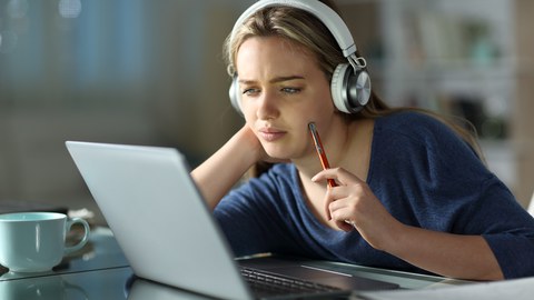  Das Foto zeigt eine junge Frau an einem Tisch. Sie schaut nachdenklich auf ihren Laptop. 