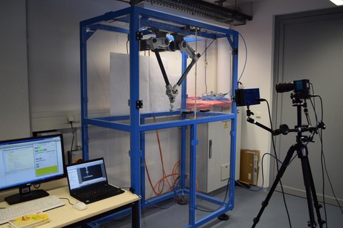 Deltaroboter mit optischem Messsystem. Neben einer Deltakinematik mit Servoantrieben ist ein High-speed Kamerasystem mit extra Beleuchtung zu sehen. Ebenso sind Rechner für Versuchsstandssteuerung und Messdatenaufzeichnung zu sehen.