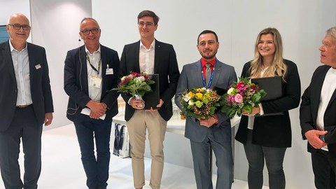 Foto von 5 Personen in Businessklediung, die in die Kamera schauen. Drei Personen halten Blumensträuße.