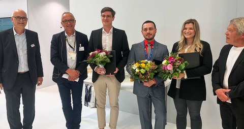 Foto von 5 Personen in Businessklediung, die in die Kamera schauen. Drei Personen halten Blumensträuße.