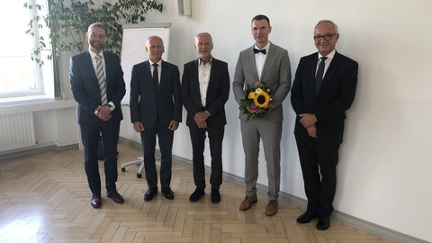 Foto von 5 Personen in Businesskleidung, die in die Kamera schauen. Eine Person hält einen Blumenstrauß.