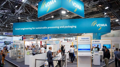 Foto eines großen Messestands. Im oberen Brecih hängen große Transparente mit der Aufschrift Smart engineering for sustainable processing and packaging und VDMA. Darunter unterhalten sich Menschan angeregt über die Exponate.