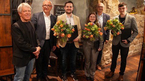 Foto einer Personengruppe in rustikalem Kellerambiente. Drei Personen haben einen Blumenstrauß.