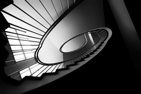 Treppe im Andreas-Schubert-Bau. Das Foto wurde vom Boden aus aufgenommen und ist in schwarz-weiß. Aus dieser Perspektive verläuft die Treppe spiralförmig nach oben.