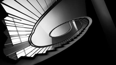 Treppe im Andreas-Schubert-Bau. Das Foto wurde vom Boden aus aufgenommen und ist in schwarz-weiß. Aus dieser Perspektive verläuft die Treppe spiralförmig nach oben.