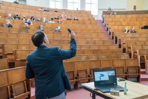 Foto eines Mannes im Anzug, der ein kleines Obejekt hochhält. Im Hintergrund sind Studierende als Zuhörer zu erkennen.