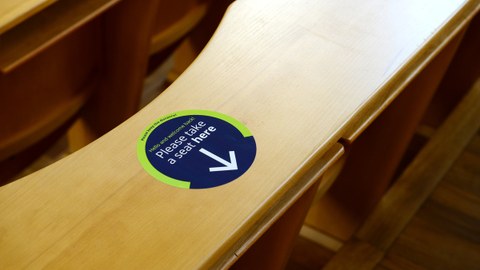 Foto eines Hörsaals mit Hinweisaufkleber an einem Sitzplatz mit der Aufschrift "Please take a seat here.". 