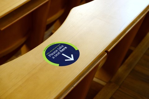 Foto eines Hörsaals mit Hinweisaufkleber an einem Sitzplatz mit der Aufschrift "Please take a seat here.". 
