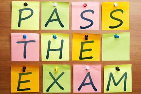 Auf dem Bild sind bunte Notizzettel auf einem Hintergrund aus Holz angeordnet. Jeder Zettel ist mit einem Großbuchstaben beschriftet. Zusammen ergeben sie die Wörter "Pass the exam".