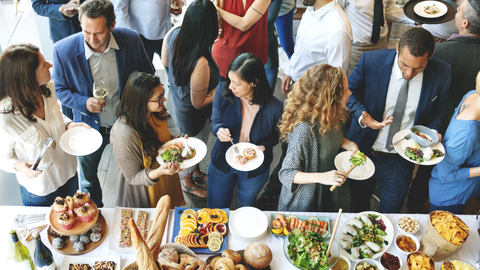 Das Foto zeigt viele Menschen in Businesskleidung. Sie stehen vor einem Buffet und reden miteinander. 