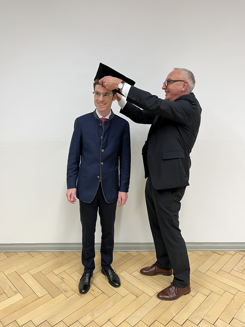 Ein Mann im schwarzen Anzug setzt einem anderen Mann einen Doktorhut auf.