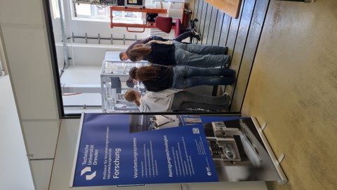 Im Vordergrund ein Plakat mit Aufschrift Forschung. Im Hintergrund 5 Mädchen an einem Versuchsstand.