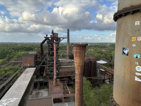 Ausblick von einem erhöhten Standpunkt auf mehrere rostige Schornsteine und Gebäude aus Metall in mitten von Bäumen und Sträuchern.