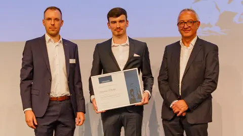 Drei Männer im Anzug. Der mittlere hält eine gerhmte Urkunde in den Händen.
