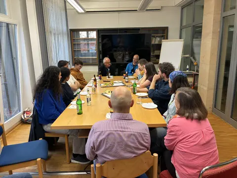 Ca. 12 Personen sitzen in Alltagskleidung an einem Koferenztisch. An den Längsseiten junge Menschan, an den Querseiten ältere Menschen.