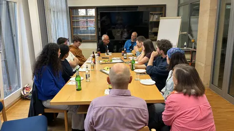 Ca. 12 Personen sitzen in Alltagskleidung an einem Koferenztisch. An den Längsseiten junge Menschan, an den Querseiten ältere Menschen.