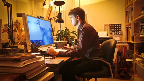 Mann sitzt in einem Büro mit Regale, Drucker und einem PC mit großem Monitor im orangen Licht.