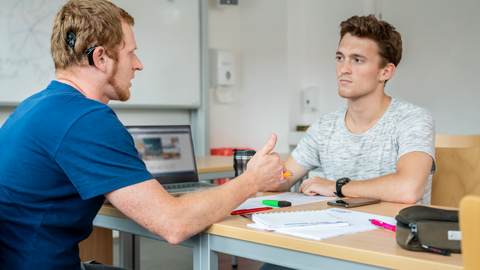 Das Foto zeigt zwei Studenten in einem Seminarraum. Sie sitzen sich gegenüber und sprechen miteinander. Zwischen ihnen liegen Zettel und Stifte. Einer der beiden Studenten trägt ein Hörgerät