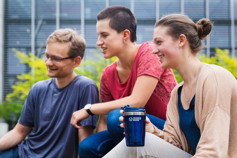 Foto: Drei Studenten sitzen beisammen. Eine davon hält einen TU Dresden Becher in der Hand.