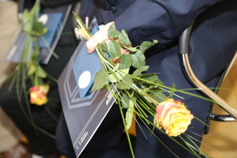 Das Foto zeigt die Hände einer Person im Anzug. Sie hält eine Rose, einen Umschlag und ein Heft in der Hand.