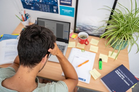 Das Foto zeigt einen Studenten an seinem Schreibtisch. Vor ihm steht sein Laptop und viele weitere Büromaterialien. Mit den Händen stützt er seinen Kopf ab. 