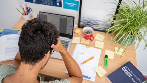 Das Foto zeigt einen Studenten an seinem Schreibtisch. Vor ihm steht sein Laptop und viele weitere Büromaterialien. Mit den Händen stützt er seinen Kopf ab. 