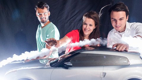 Foto: 3 Studierende blicken begeistert auf ein Modellauto, das von einer sichtbaren Luftströmung umflossen wird. 