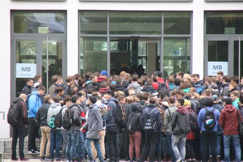 Foto vieler Studierender, die vor einer großen Eingangstür stehen. Es scheint so, als sei der Eingang zu schmal für die vielen Studierenden.