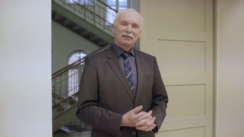 Foto: Der Dekan der Fakultät, Prof. Stelzer steht in einer Tür und spricht in Richtung der Kamera. Im Hintergrund ist ein Treppenhaus zu sehen.