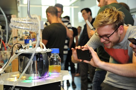 Foto: Ein Student fotografiert während einer Ausstellung mit seinem Handy den Bioreaktor der TU-dresden, er blickt begeistert