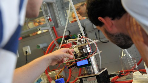 Foto: Zwei Studenten stehen vor dem kleinen Bioreaktor des Studierenden-Teams der Universität Stuttgart und betrachten ihn genau.