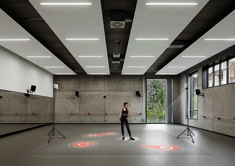 Foto: Eine Tänzerin steht in einem großen Raum, sie trägt einen Gürtel und Armbänder, die taktile Vibrationssignale aussenden. Auf den Boden werden mehrere rote Felder projiziert.