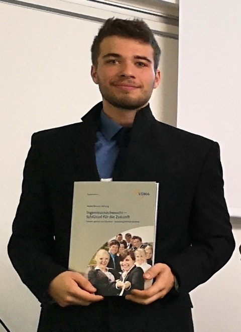 Foto: Der Student Eric Otta erhält den Kreativitätspreis des Deutschen Textilmaschinenbaues 2018. Der Preisträger hält ein Heft in der Hand und lächelt in die Kamera. 