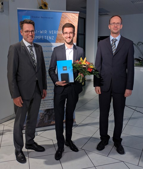 Drei Männer in dunklen Anzügen stehen nebeneinander. Der jüngste Mann in der Mitte hält eine Urkunde und einen Blumenstrauß in der Hand.