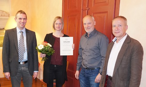 Auf dem Foto stehen vier Personen nebeneinander. Die zweite Person von links ist eine junge Frau. Lydia Hofmann hält in ihrer linken Hand eine Urkunde und in ihrer rechten Hand einen Blumenstrauß. Sie hat glatte, fast schulterlange blonde Haare. 