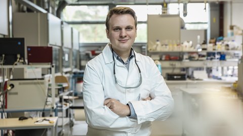 Das Foto zeigt Josua Viethen, der einen weißen Kittel trägt, die Arme vor der Brust verschränkt und in einem Labor steht.