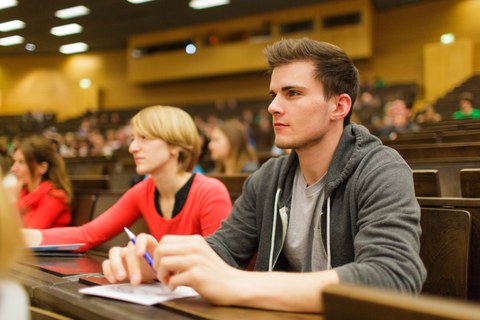 Das Foto zeigt Studierende im Hörsaal. Im rechten Bildvordergrund sieht man einen Student mit kurzen Haaren und grauer Jacke, die Arme stützt er auf dem Tisch ab. Neben ihm sitzt eine blonde Studentin mit rotem Shirt.