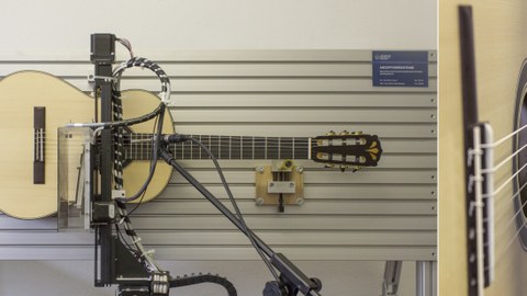 Das Foto ist zweigeteilt. Der überwiegende linke Teil zeigt eine Akustikgitarre, die in eine Wandvorrichtung gespannt ist. Der Anzupftest untersucht wie lange es dauert, bis ein Ton hörbar ist und wie lange der Ton hörbar bleibt. Der kleine rechte Teil des Fotos zeigt eine Detailaufnahme der Gitarrensaiten, die gerade von der Vorrichtung angezupft werden.