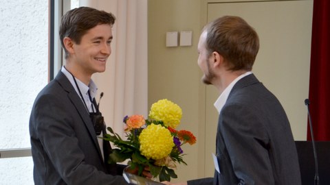 Das Foto zeigt, wie ein junger Mann im Anzug gerade einen Blumenstrauß an Mauritz Mälzer übergibt. Er trägt ebenfalls einen dunklen Anzug und freut sich sichtlich über den Preis. 