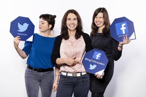 Drei junge Frauen stehen nebeneinander vor einem weißen Hintergrund. Alle drei halten ein achteckiges Schild in der Hand. Auf den Schildern steht die Webadresse der Social-Media-Kanäle "@ingTUDresden".